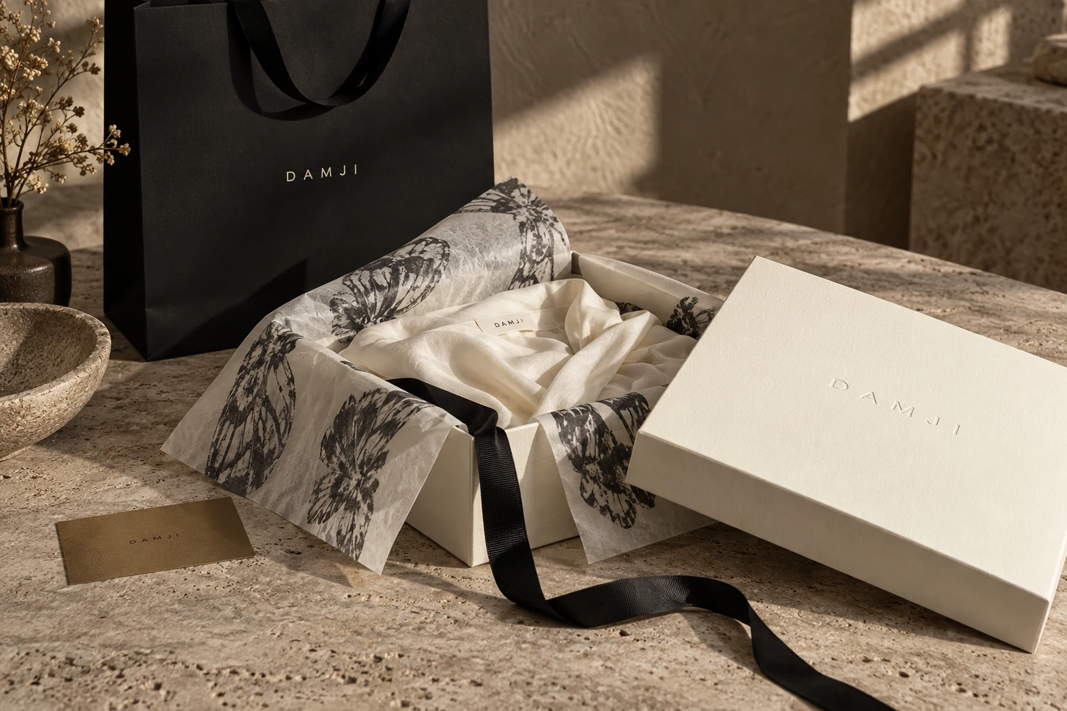 Mockup da experiência de embalagem DAMJI: caixa marfim, papel estampado e sacola preta